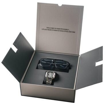 Metal wristwatch in a black open presentation box, with a gray sleeve behind and visible text on it.