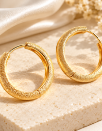 Textured gold hoop earrings resting on a light stone surface with soft ivory fabric in the background  informative image describing the jewelry setup