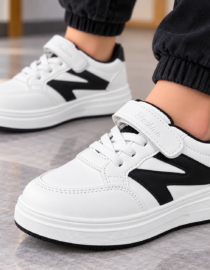 White sneakers with black side panels and a Velcro strap worn with black joggers on a tiled floor