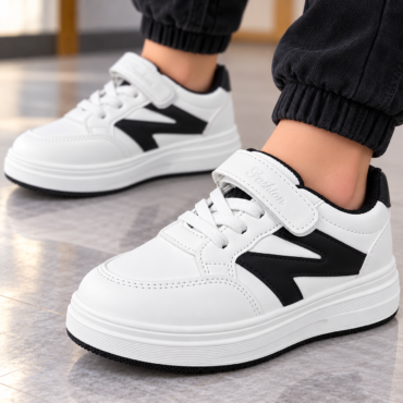 White sneakers with black side panels and a Velcro strap, worn with black joggers on a tiled floor.