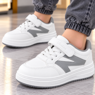 Close-up of white kids' sneakers with gray panels and a velcro strap, worn with cuffed jeans on a tiled floor