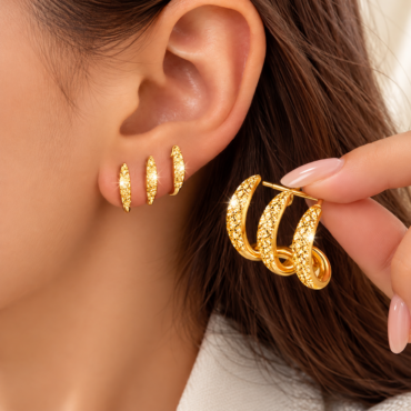 Close-up of a woman's ear adorned with several gold, diamond-studded hoop earrings, held by a hand showing additional hoops nearby.