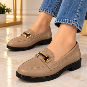 Beige leather loafers with gold buckle hardware, worn with blue jeans on a marble floor indoors.