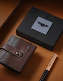 Brown leather wallet with gray card slots and a snap strap on a tan surface with a black gift box in the background